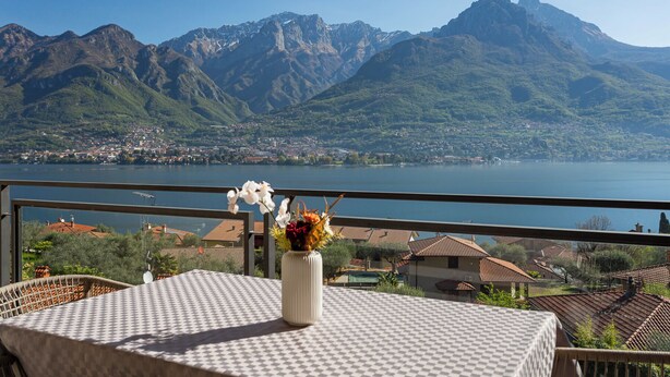 Een balkon met een tafel, een vaas met bloemen en uitzicht op bergen en een meer.