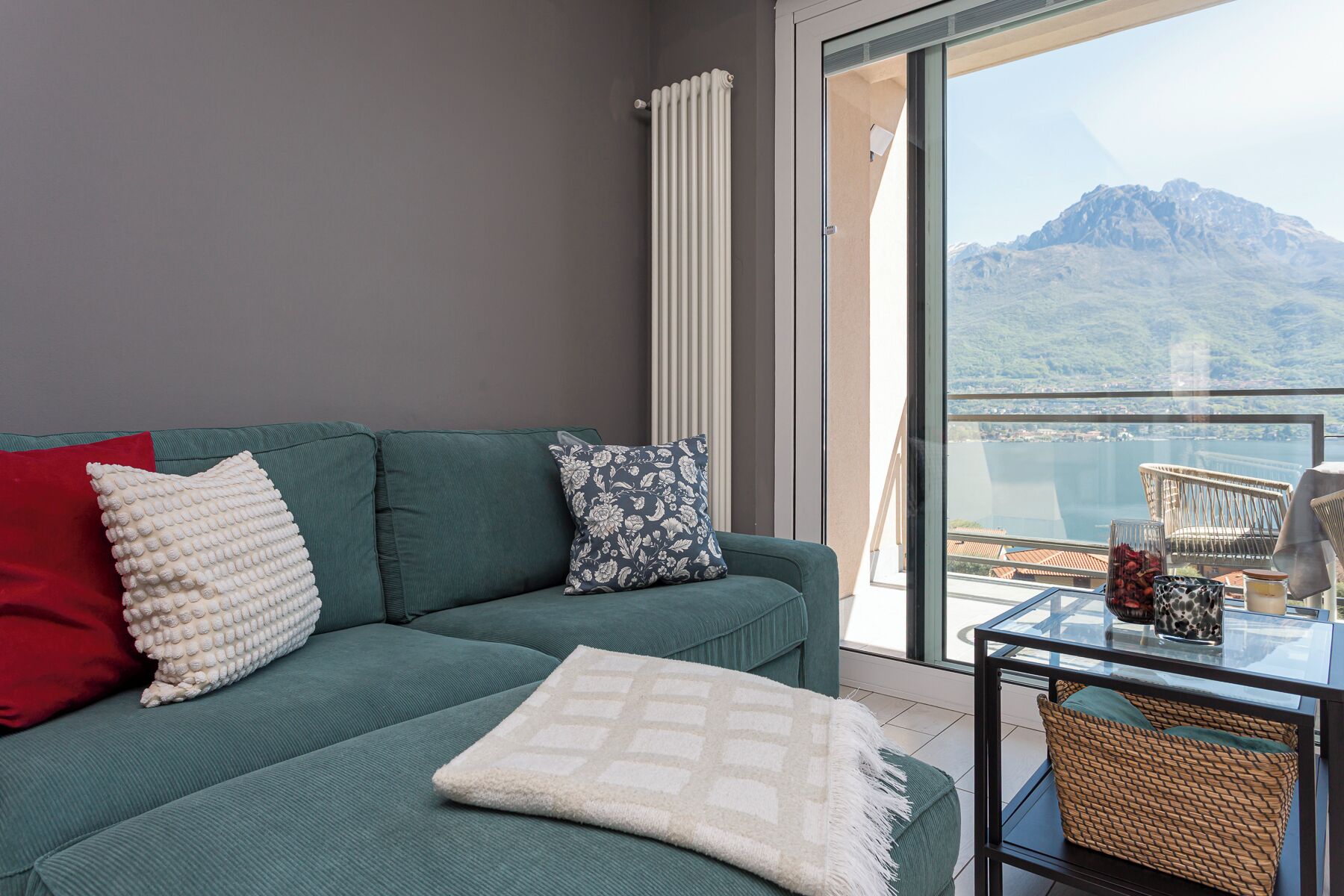 Een woonkamer met een groene bank, een rode kussen, een gestreept kussen, een witte plaid en een glazen tafel met rieten manden.