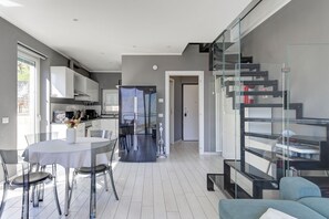 A modern kitchen with white cabinets, a dining area with a glass table and chairs, and a staircase with glass railings. - Open View Apartment by Wonderful Italy (Onno)