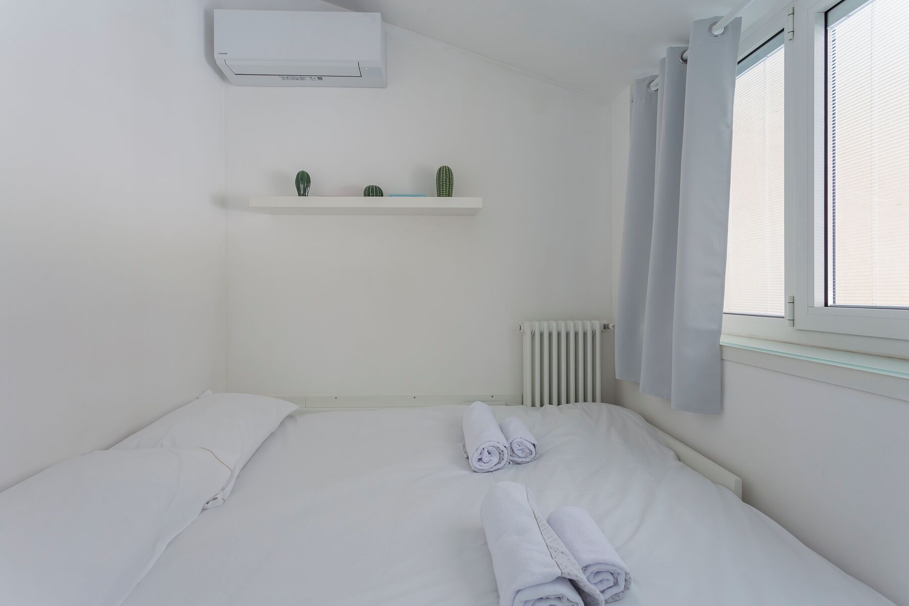 Habitación de hotel con cama, radiador, ventana con cortinas y aire acondicionado instalado en la pared.