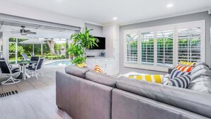 A modern living room with a grey sofa, a dining area with a table and chairs, a TV, and a large window with blinds.