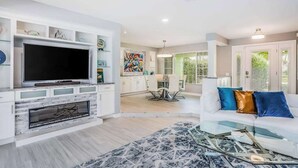 A modern living room with a flat-screen TV, a fireplace, a dining area, and a kitchen in the background.
