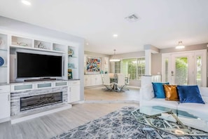 A modern living room with a flat-screen TV, a fireplace, a dining area, and a kitchen in the background. - Waterfront escape in the heart of the action with pool, spa, grill, & WiFi (Cape Coral)