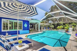 A pool area with lounge chairs, a covered seating area, and a building with a blue exterior. - Waterfront escape in the heart of the action with pool, spa, grill, & WiFi (Cape Coral)