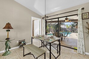 A dining area with a table set for four, a bench, a decorative lamp, and a bookshelf with books. - Cape Escape - Dog-Friendly | Pool, Bar & Fenced Yard (Cape Coral)