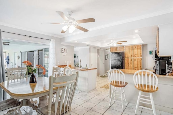 A modern kitchen with wooden cabinets, a central island, and a dining area with white chairs. - Snowbird-friendly home with enclosed yard, pool, patio, AC, & WiFi - 2 dogs OK (Cape Coral)