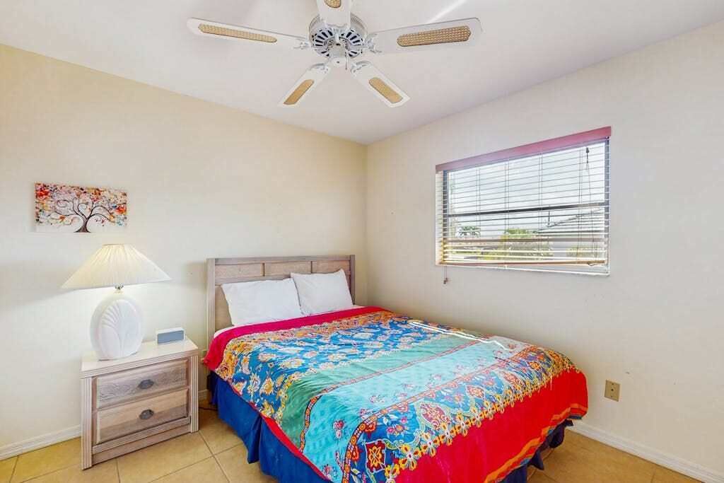 Un dormitorio con cama, mesita de noche, lámpara, ventilador de techo, ventana con persianas y una obra de arte colgada en la pared.