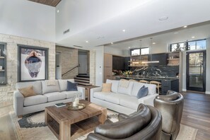 A spacious living area with a sofa, coffee table, and leather chairs, a kitchen with a marble countertop, and a staircase leading to another level. - Chateau Cortina-Newly Built 2026, 5 Guest Suites, Ski-In/Out, Ski Lounge, A/C (Mountain Village)