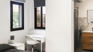 A bedroom with a bed, a desk, a chair, a bedside table, a window, and a bathroom with a mirror and sink.