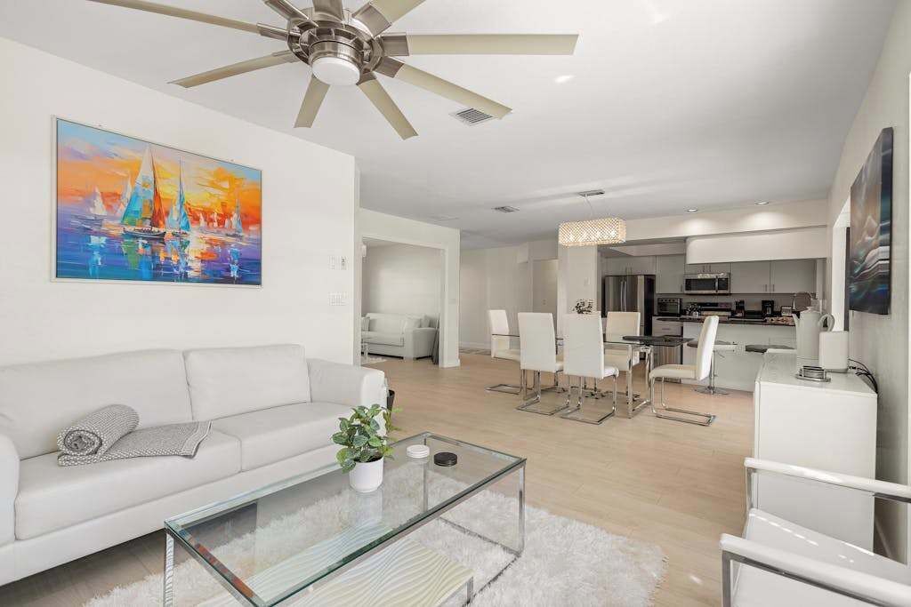 A modern living room with a white sofa, a glass coffee table, a ceiling fan, and a painting of sailboats.