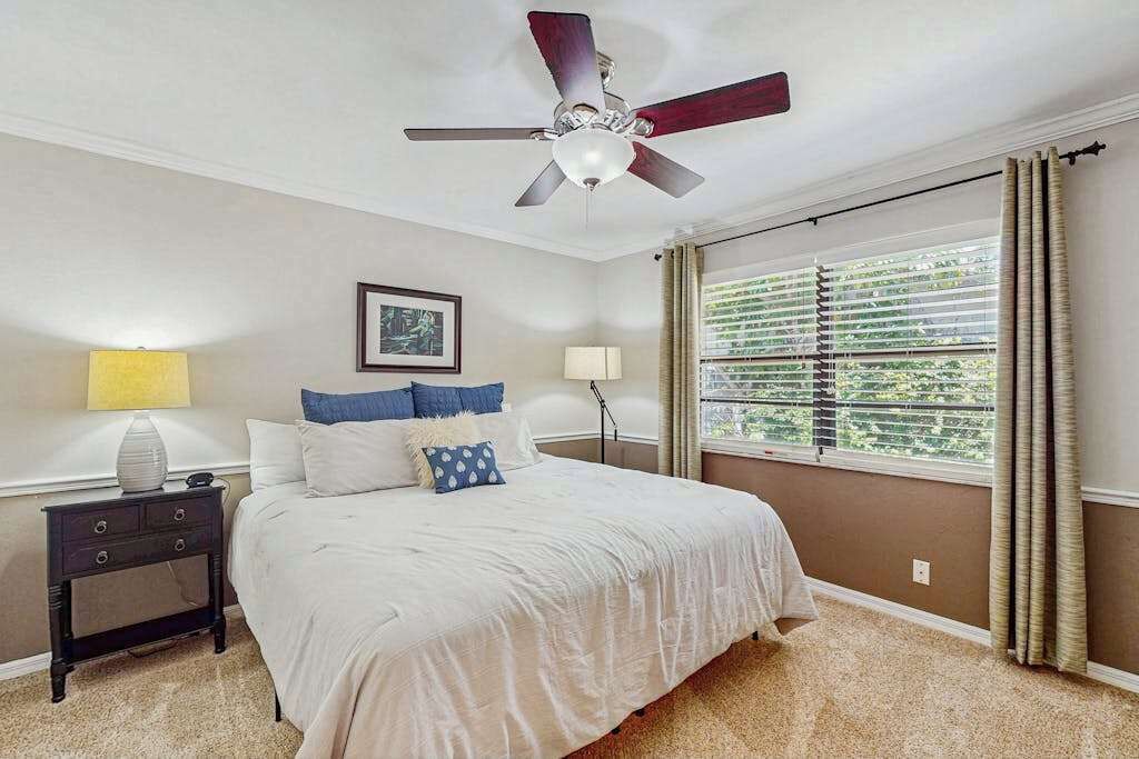 A bedroom with a bed, nightstand, ceiling fan, window with blinds, and a picture on the wall.