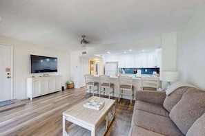 A modern living room with a sofa, a coffee table, a TV, and a kitchen area with white cabinets and a tiled backsplash. - Beachfront condo with pool, private balcony, and washer and dryer (Englewood)