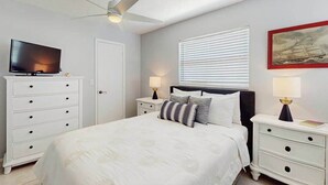 Un dormitorio con cama, televisor, ventilador de techo, ventana con persianas y un cuadro en la pared.