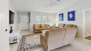 En stue med en beige sofa, et sofabord og et tæppe med et mønster. To billeder hænger på væggen, der er en loftvifte, og en dør der fører til et andet rum.