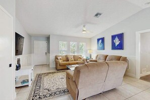 A living room with a beige sofa, a coffee table, and a patterned rug. There are two framed pictures on the wall, a ceiling fan, and a door leading to another room. - Snowbird-friendly home with private pool, water views, patio & sunroom (Englewood)