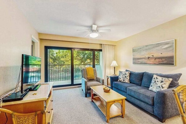A living room with a sofa, coffee table, TV, and a balcony with a view. - Spacious, waterfront condo with outdoor pool, tennis, & volleyball (Naples)