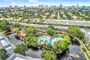 An aerial view of a resort with a swimming pool surrounded by lush greenery, a small bridge, and a view of the city skyline in the background. - Spacious, waterfront condo with outdoor pool, tennis, & volleyball (Naples)