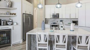 Cocina moderna con armarios blancos, una isla central con taburetes de bar, electrodomésticos de acero inoxidable e iluminación colgante.