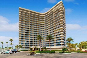 A multi-story hotel with a curved facade, balconies, and a parking area with palm trees. - Waterview condo with shared pool, pickleball, and WIFI (Marco Island)