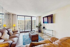 A modern living room with a leather sofa, a coffee table, and a large window offering a city view. - Waterview condo with shared pool, pickleball, and WIFI (Marco Island)