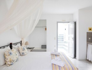 Quarto com cama, mesa de cabeceira, luminária, janela com cortinas e banheiro visível por uma porta aberta.