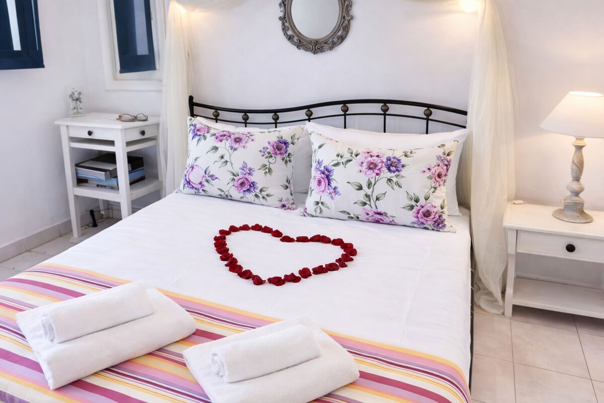 A neatly made bed with floral pillows, a heart-shaped decoration of red roses, and folded white towels.
