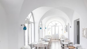 A bright, white interior with arched ceilings and a central open door leading to another room. Tables and chairs are arranged along the walls, and there are blue hanging lamps. - Hotiday Santorini Sea View (Santorini)