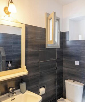 Une salle de bain avec des carreaux foncés, un lavabo blanc, un miroir avec un éclairage intégré et des toilettes.