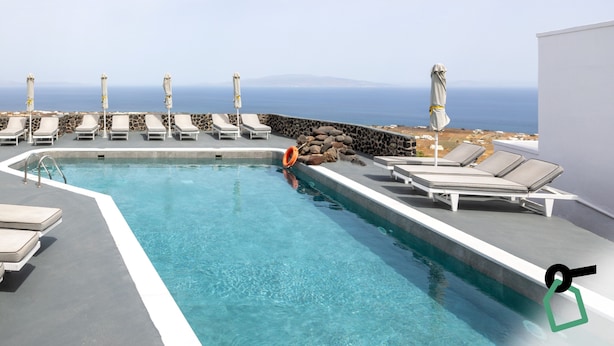 A pool with lounge chairs and umbrellas, a stone wall, and a view of the ocean. - Hotiday Santorini Sea View (Santorini)