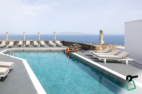 A pool with lounge chairs and umbrellas, a stone wall, and a view of the ocean.