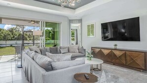 A modern living room with a sectional sofa, a coffee table, a TV, and a balcony with a view of greenery. - Contemporary Getaway with an Enclosed Yard, Fast WiFi, & Furnished Lanai (Bonita Springs)