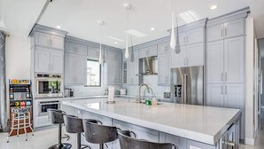 A modern kitchen with grey cabinets, a large island with bar stools, stainless steel appliances, and pendant lighting.