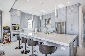 A modern kitchen with grey cabinets, a large island with bar stools, stainless steel appliances, and pendant lighting. - Spacious modern home close to beach with pool, central AC, & washer/dryer (Bonita Springs)