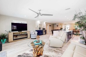 Ein modernes Wohnzimmer mit Flachbildfernseher, einem Glastisch, Deckenventilator und einer Küche im Hintergrund.