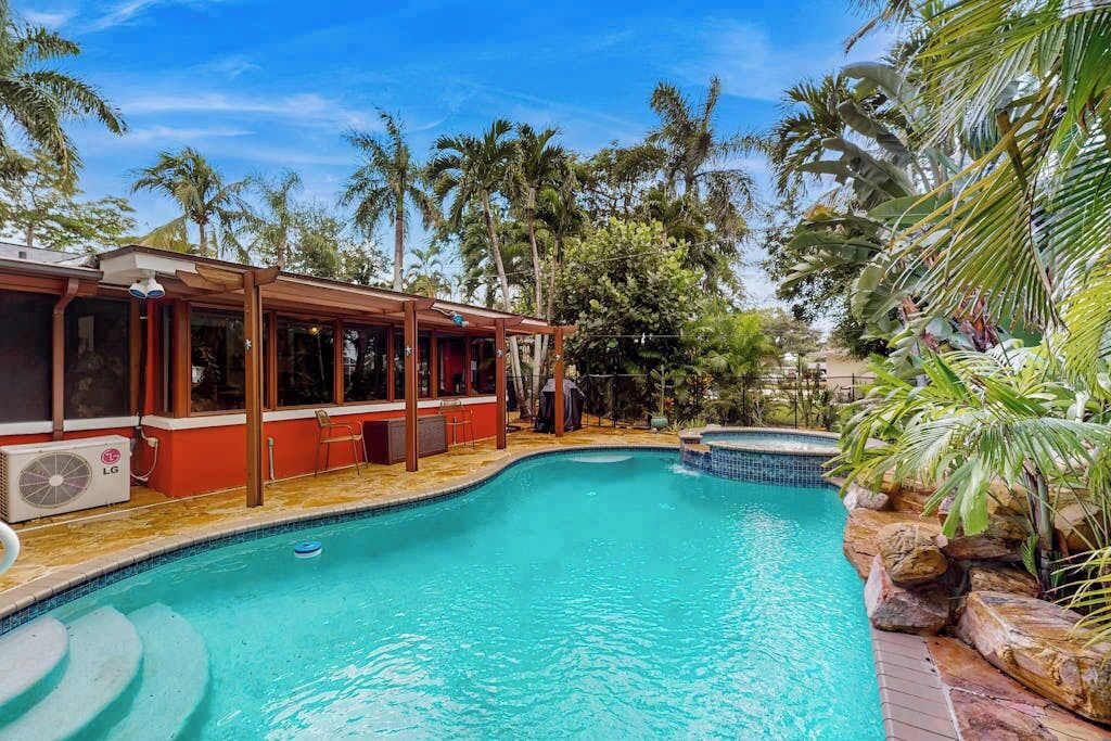 A swimming pool with a wooden deck, a covered seating area, and a view of palm trees.