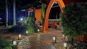 A pathway leading to a building with an archway, surrounded by greenery and outdoor lighting. - Bali-inspired coastal home w/private pool, spa, and tiki room (Cape Coral)