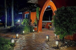 Un chemin menant à un bâtiment doté d’une arche, entouré de verdure et éclairé par des lampes extérieures.