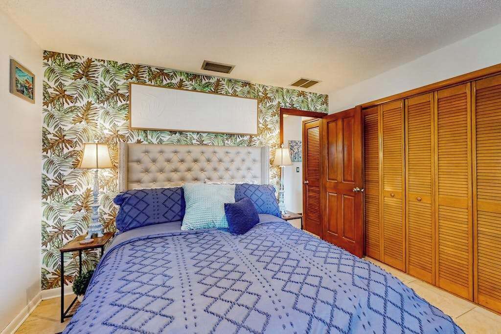 A bedroom with a tufted headboard, a large bed with patterned bedding, two bedside lamps, a wooden wardrobe, and a tropical leaf-patterned wallpaper.