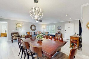 Ein Essbereich mit einem großen Holztisch, Stühlen, einer Chandelierleuchtre und einer Couch im Hintergrund.