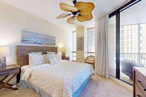 A hotel room with a large bed, a ceiling fan, a view of the city through large windows, and a wicker headboard. - Stylish Water-View Condo w/ Balcony, Multiple Pools & Tennis Courts (Cape Coral)