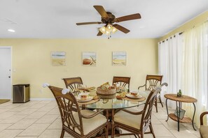 A dining area with a glass table surrounded by wicker chairs, a ceiling fan, and wall art depicting beach scenes. - Open-Air Waterfront Retreat | Private Pool (Cape Coral)