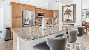 A modern kitchen with granite countertops, stainless steel appliances, wooden cabinets, and a large island with bar stools. - Stunning waterfront snowbird escape with private saltwater pool, dock, AC & W/D (Cape Coral)