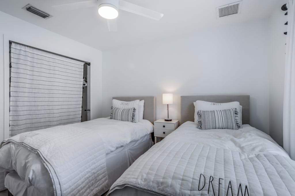 Un dormitorio con dos camas, un ventilador de techo, una mesita de noche con lámpara y una ventana con persianas.
