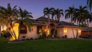 A residential house with a well-maintained lawn, palm trees, and a flagpole. - Canal-Front Home with Private Pool and Dock, WiFi, and Pool Table (Cape Coral)