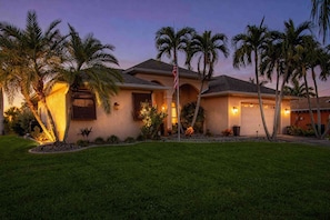 A residential house with a well-maintained lawn, palm trees, and a flagpole. - Canal-Front Home with Private Pool and Dock, WiFi, and Pool Table (Cape Coral)