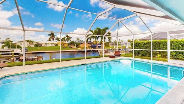 Una piscina con techo transparente, rodeada de sillones de descanso y vistas a palmeras y una masa de agua.