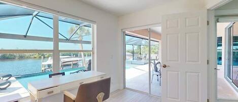 Una habitación con escritorio, silla, ventanas amplias y una puerta que da a un balcón con vista a la piscina.
