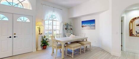 Een eethoek met een witte tafel en stoelen, een potplant en een schilderij aan de muur.