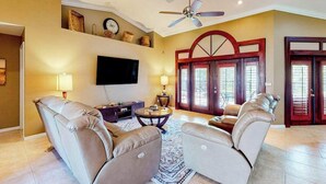 A living room with a ceiling fan, a flat-screen TV, leather sofas, a coffee table, and a patterned rug. - Inviting home with pool, water view, enclosed yard, central AC, W/D (Cape Coral)