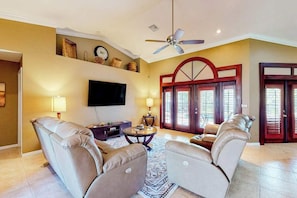 A living room with a ceiling fan, a flat-screen TV, leather sofas, a coffee table, and a patterned rug. - Inviting home with pool, water view, enclosed yard, central AC, W/D (Cape Coral)